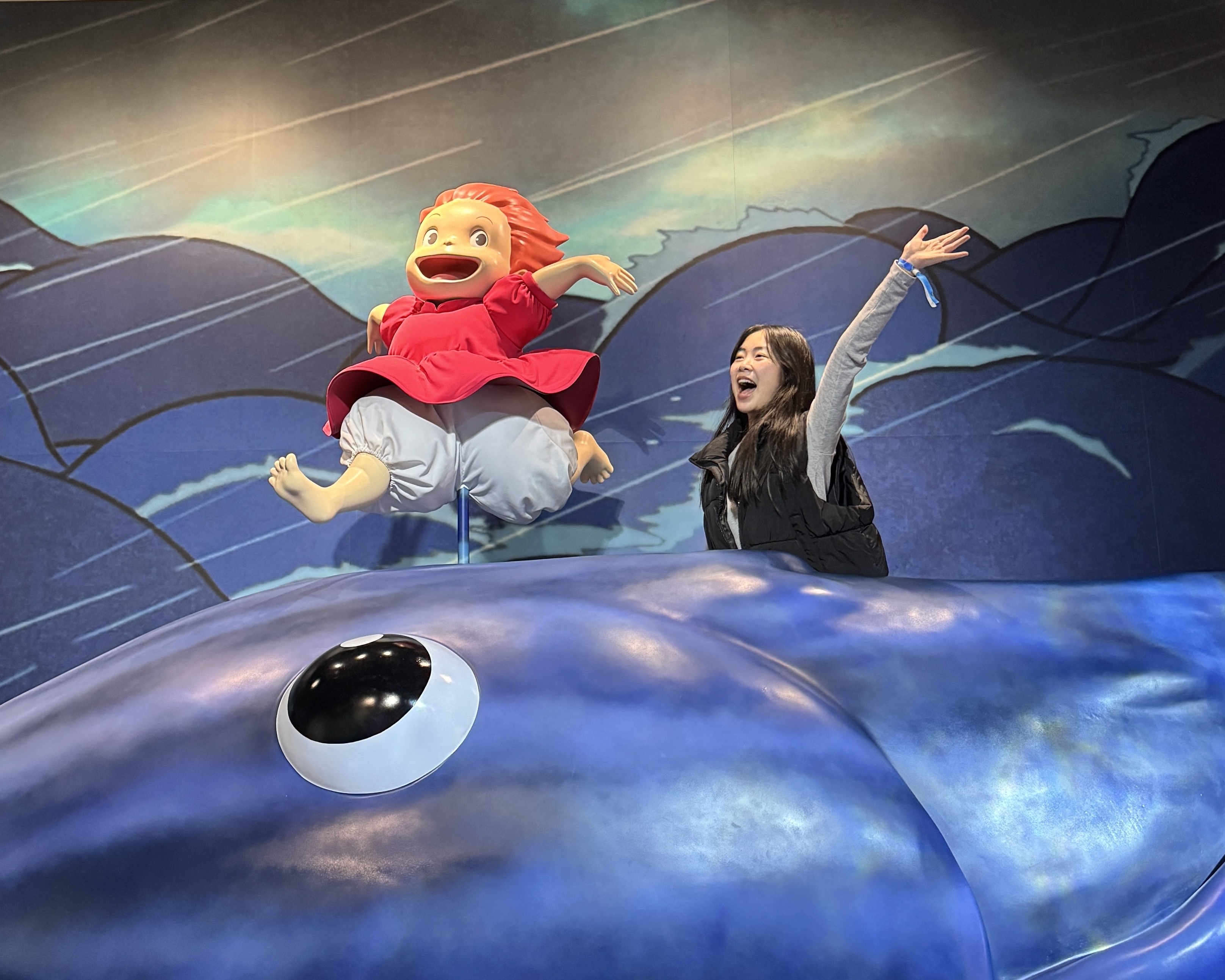 A joyful scene with a girl enthusiastically posing beside a vibrant statue of a character in red. Both are on a giant blue creature's back. Vibrant and dynamic.