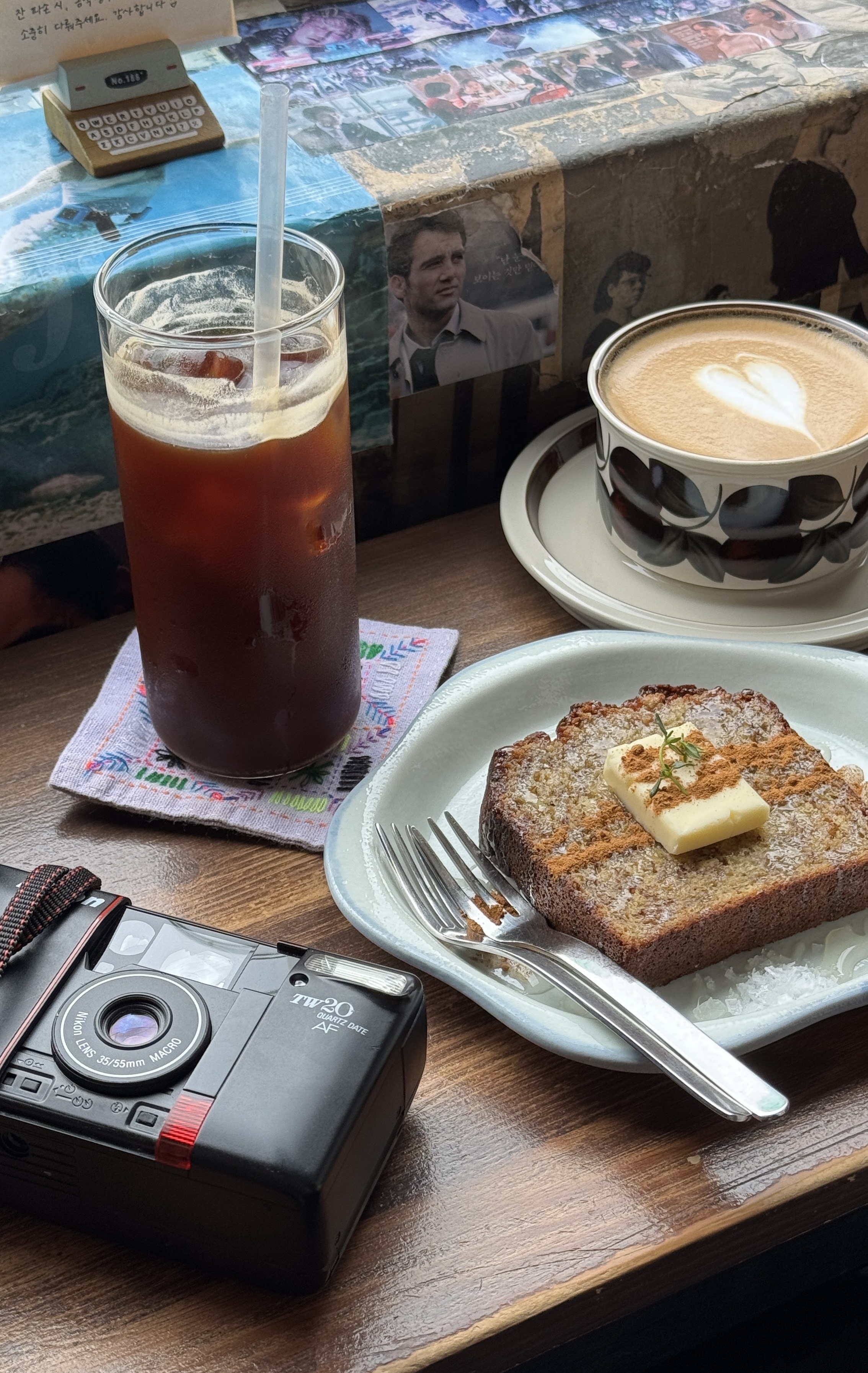 A cozy café table with iced coffee, a latte with heart foam, and banana bread topped with butter. A film camera rests nearby, creating a nostalgic vibe.