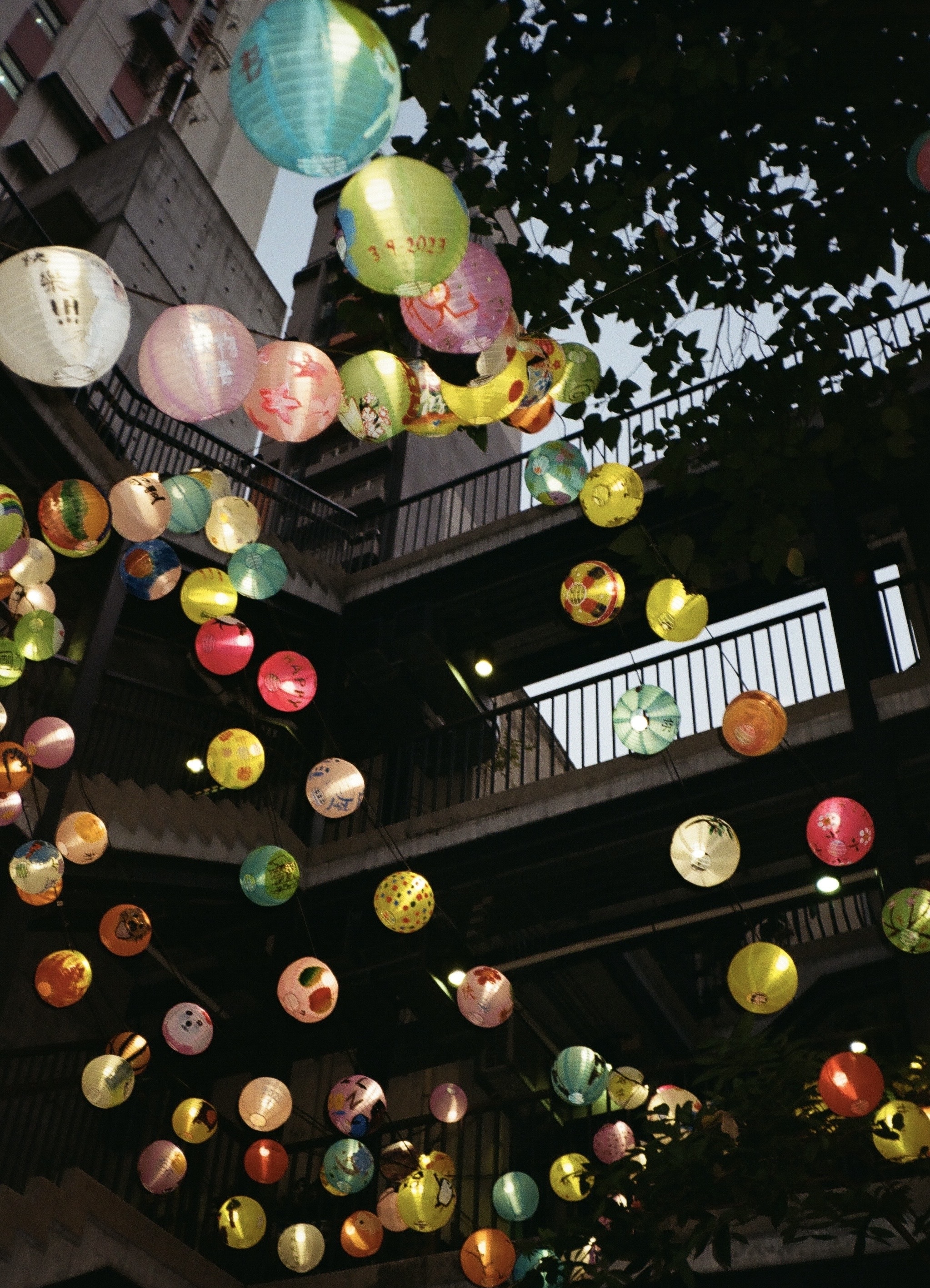 Colorful paper lanterns are strung across a courtyard at dusk, casting a warm, festive glow. Overlapping branches and balconies add depth and urban charm.