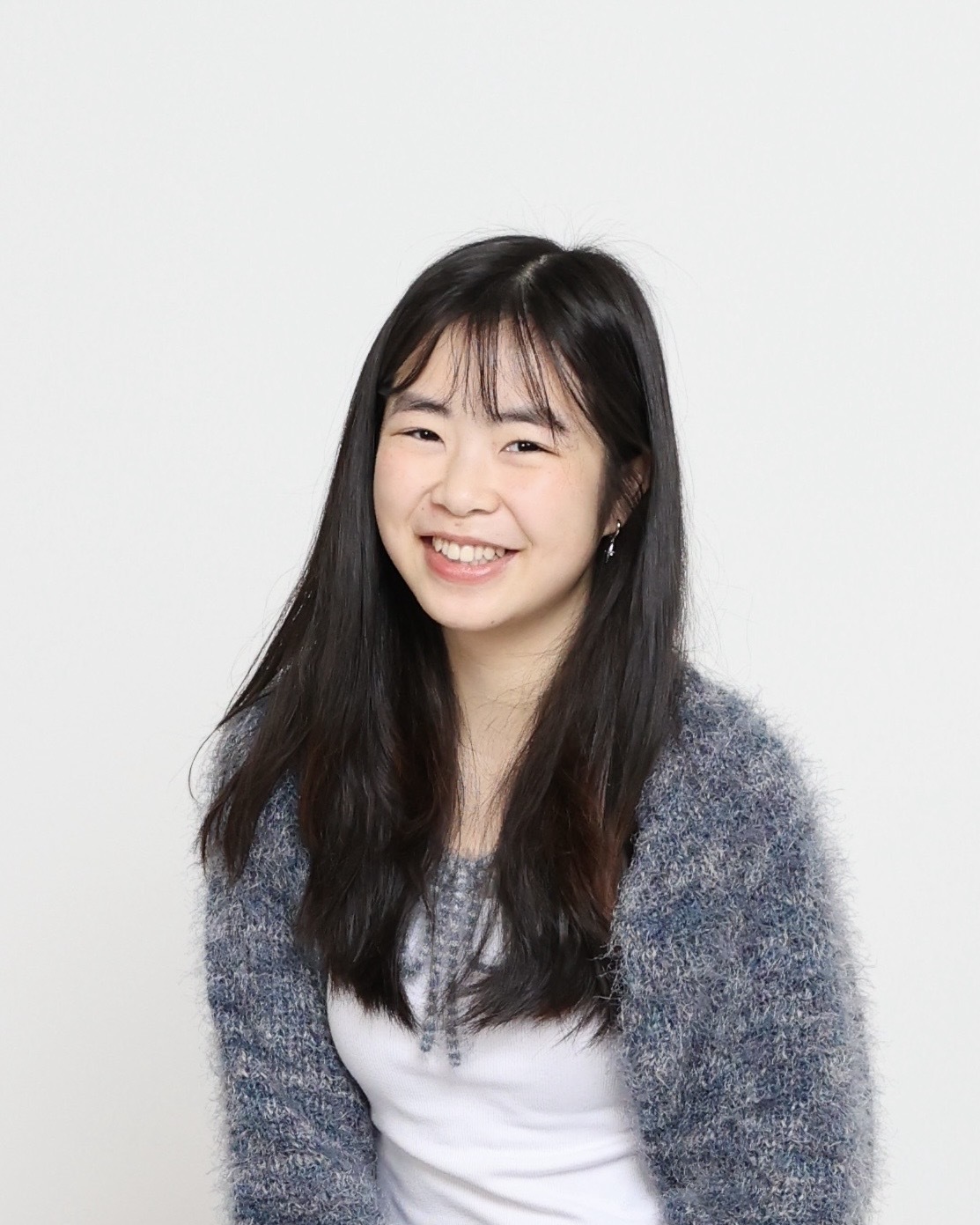 Smiling young woman with long black hair, wearing a fluffy blue cardigan and white top, sits against a plain white background. The tone is cheerful and warm.